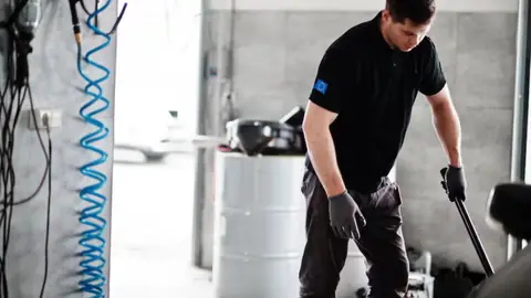 Worker in gloves using a long-handled tool on a floor beside a barrel